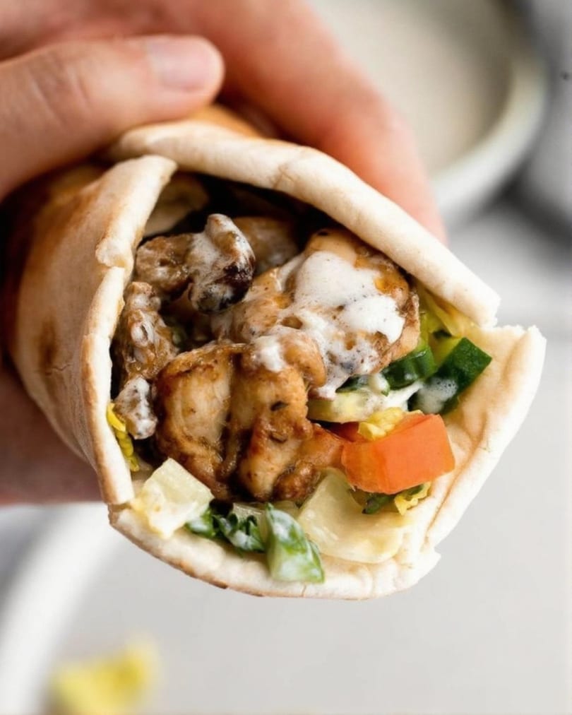 Hand holding a chicken shawarma wrap filled with meat, vegetables, and white sauce.