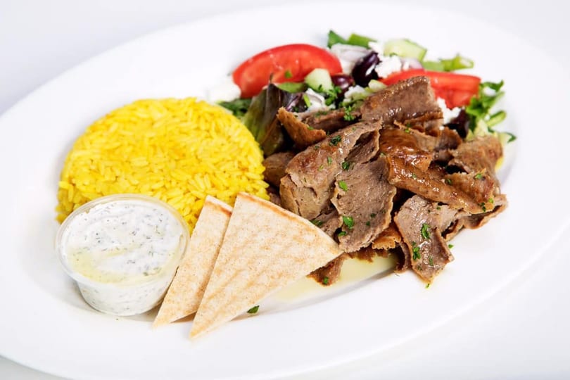 A gyro plate with sliced meat, yellow rice, pita bread, tzatziki sauce, and a side salad.