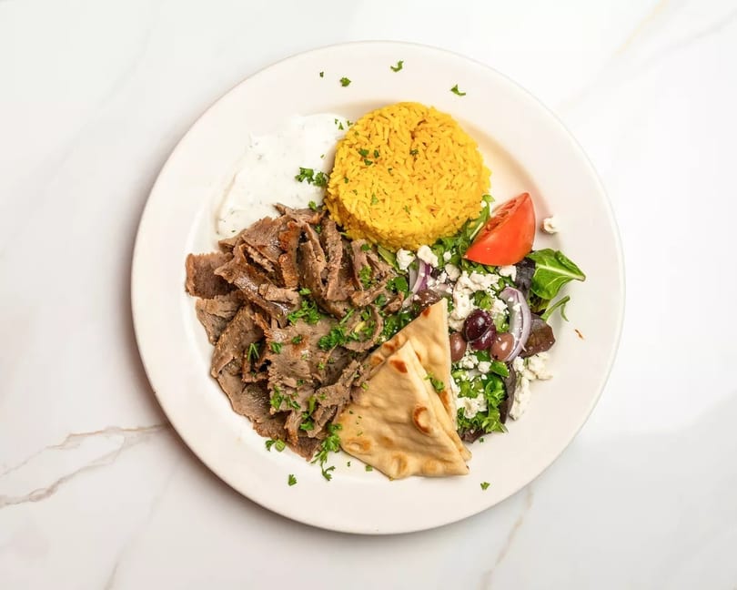 A gyro platter with sliced meat, yellow rice, Greek salad, pita bread, and tzatziki sauce.