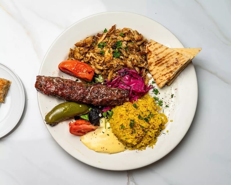 A white plate with chicken shawarma, kebab, yellow rice, grilled vegetables, and pita bread.