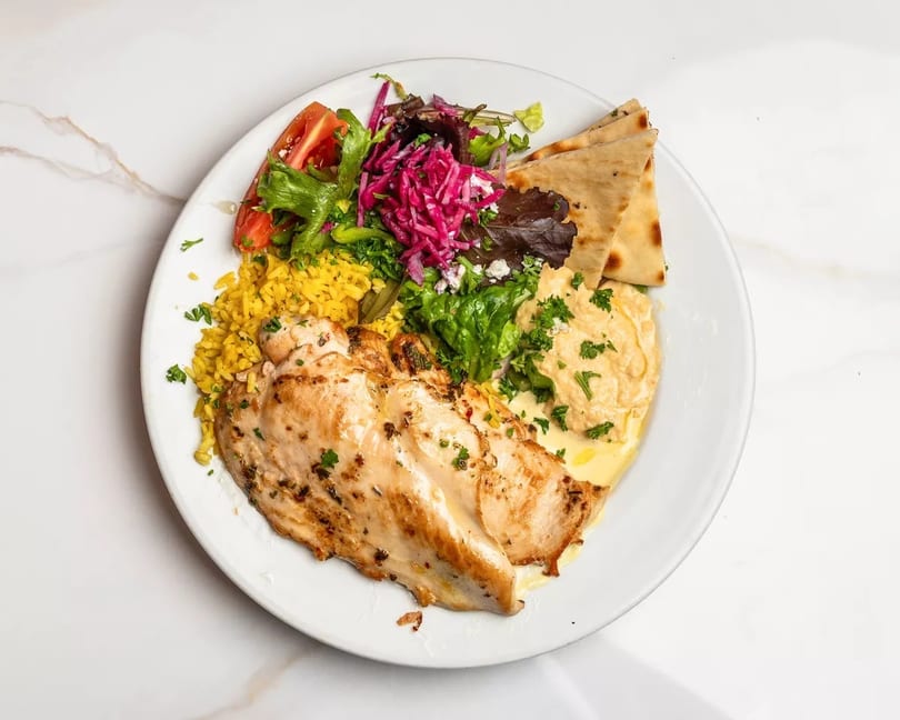 Grilled chicken breast with yellow rice, hummus, pita bread, and a fresh salad.