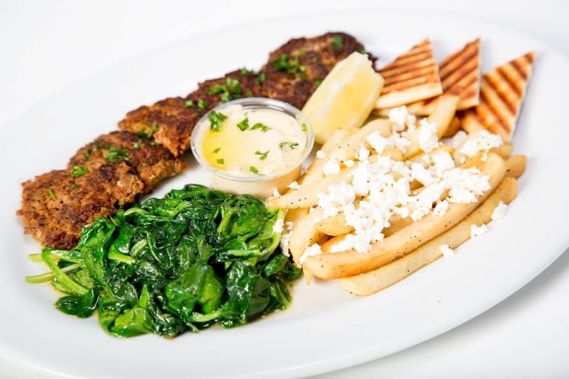 A platter with grilled ground meat, sautéed spinach, feta fries, hummus, lemon, and pita bread.